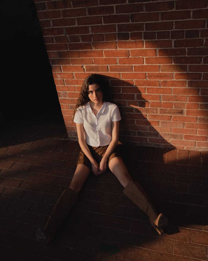 Stylish woman in white shirt and boots sitting against a brick wall in cityscape light.